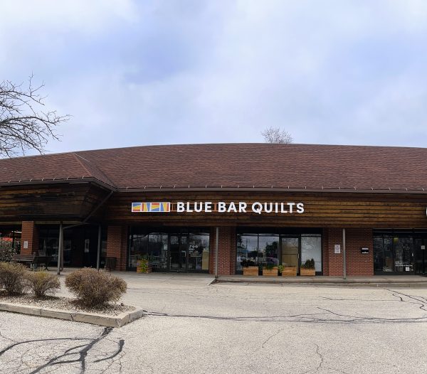 Front view of a building with a wooden facade and a sign reading "Blue Bar Quilts" above the entrance. There are potted plants near the doors.