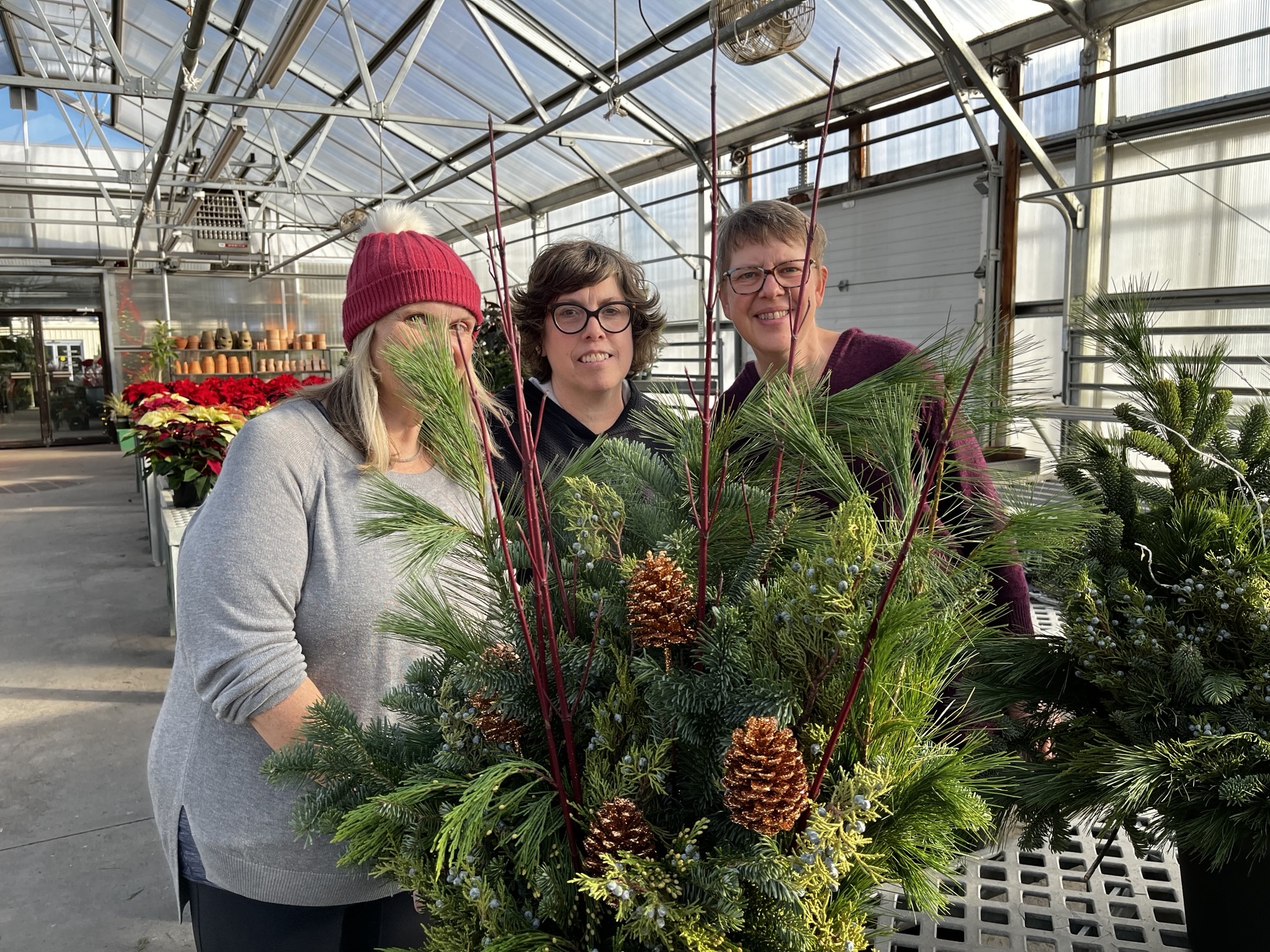 Three people in a greenhouse stand behind a large arrangement of pine branches and pine cones.