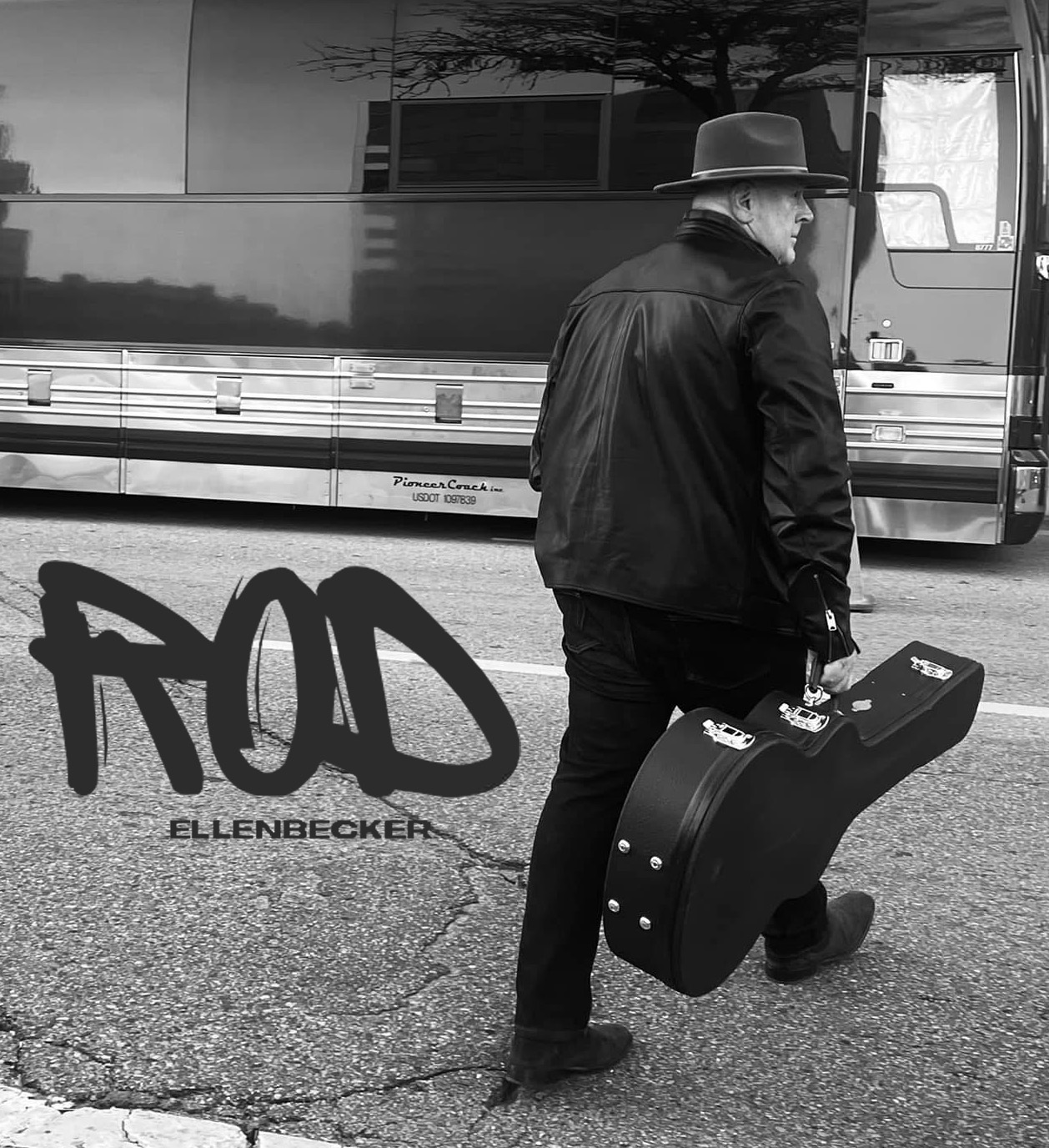 A person in a hat walks down a street beside a tour bus, carrying a guitar case.