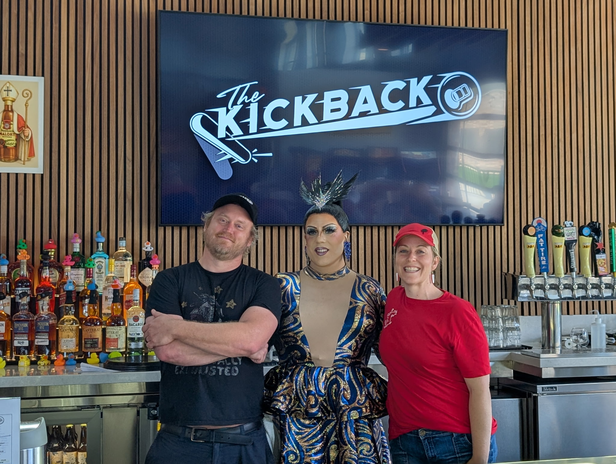 Three people pose behind a bar with liquor bottles and beer taps, standing in front of a sign that reads "The Kickback." The person in the center wears a colorful outfit and dramatic headpiece.