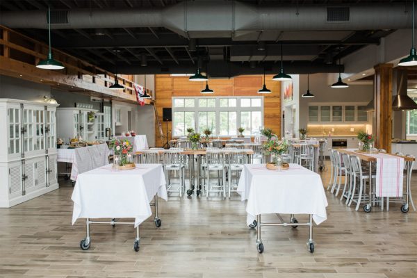 A spacious, modern event venue with tables covered in white cloths, flower centerpieces, wooden floors, and natural light from large windows creates an inviting dining atmosphere.