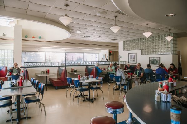 People sit at booths and tables in a retro-style diner with red and blue seating, chrome accents, and large windows, enjoying classic fare like pie.