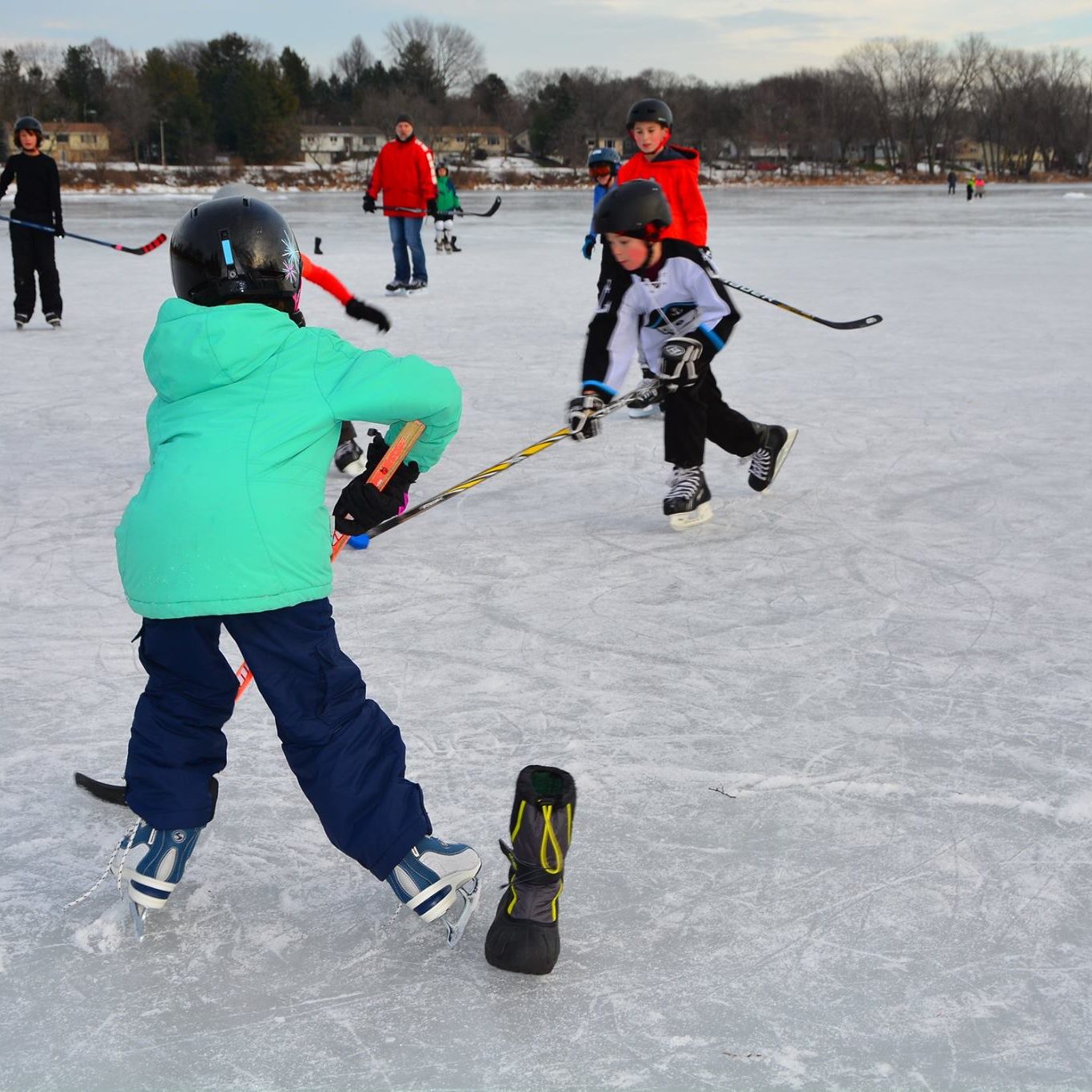 Children and adults playing ice hockey on a frozen lake, wearing helmets and winter clothing, with trees and houses visible in the background.