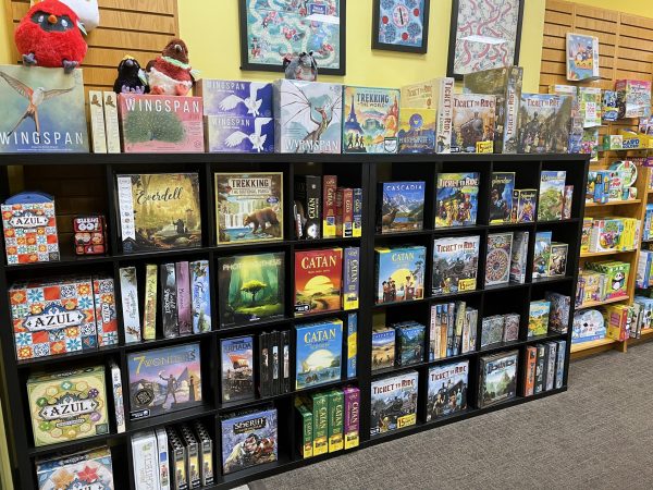 A display shelf filled with various board games, including Catan, Azul, and Wingspan, against a yellow wall with framed maps hanging above.