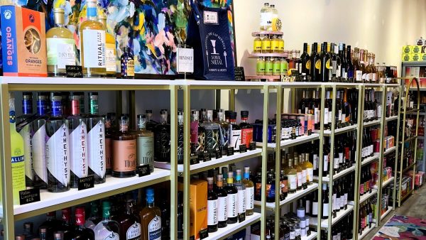 A well-stocked liquor store displays various bottles on neatly organized shelves. A colorful painting and vibrant rug add a decorative touch to the space.
