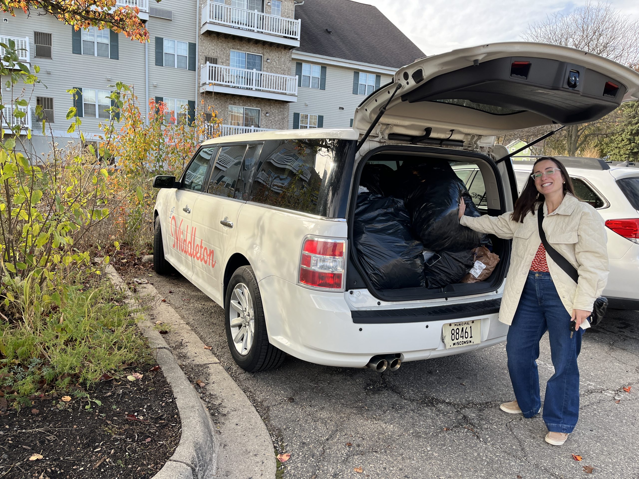 A woman stands by an open SUV trunk filled with large black garbage bags in a parking lot next to an apartment building.