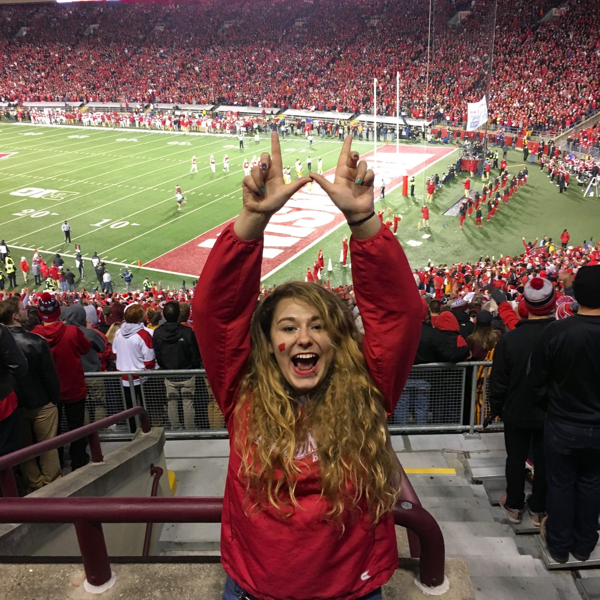 A woman in a red sweatshirt stands in a stadium, smiling and raising her hands in a "W" shape, celebrating UW Badger football with the crowd and game action visible in the background.