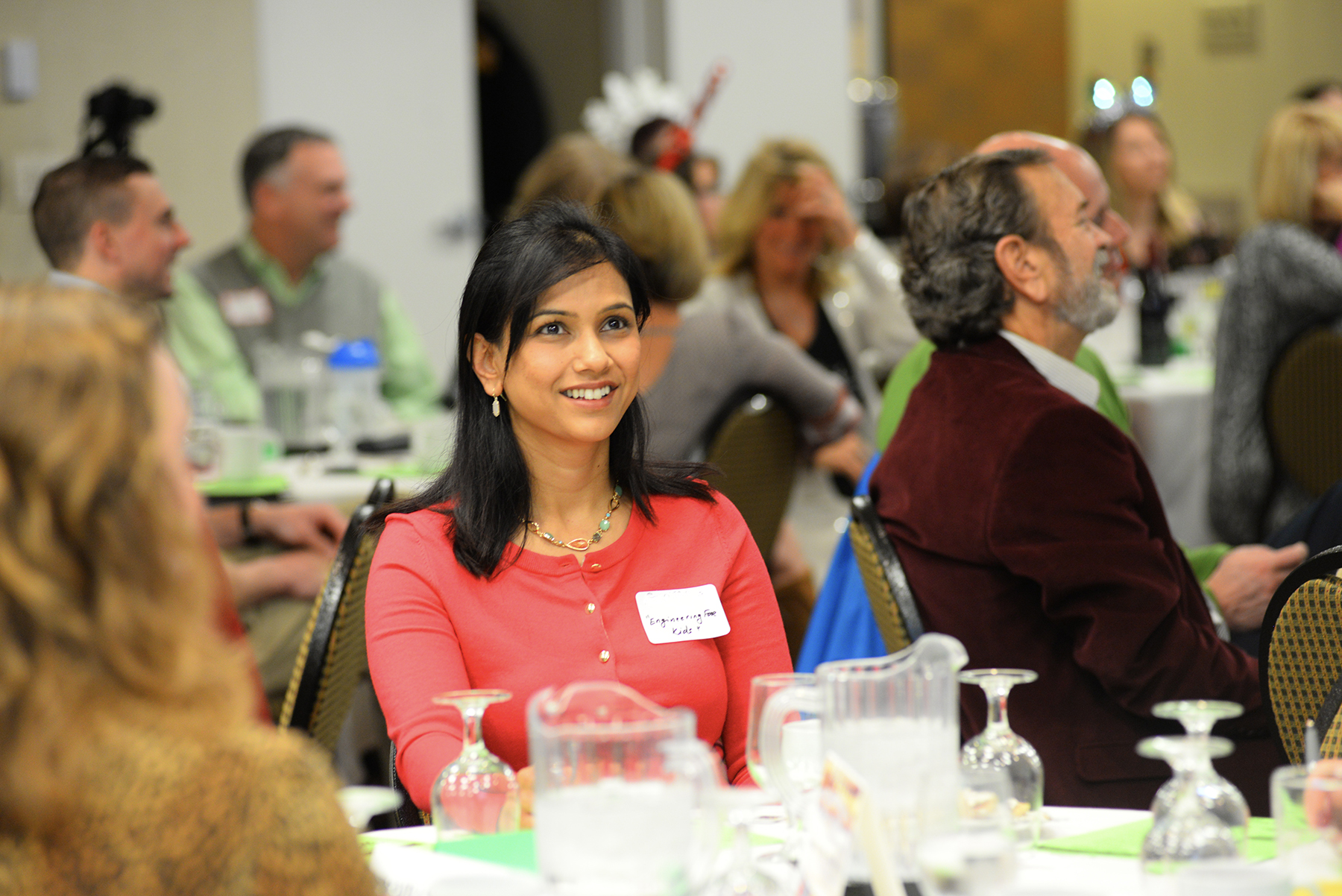 A woman in a red sweater sits at a table during a social gathering, smiling. Attendees are seated around tables in a brightly lit room.