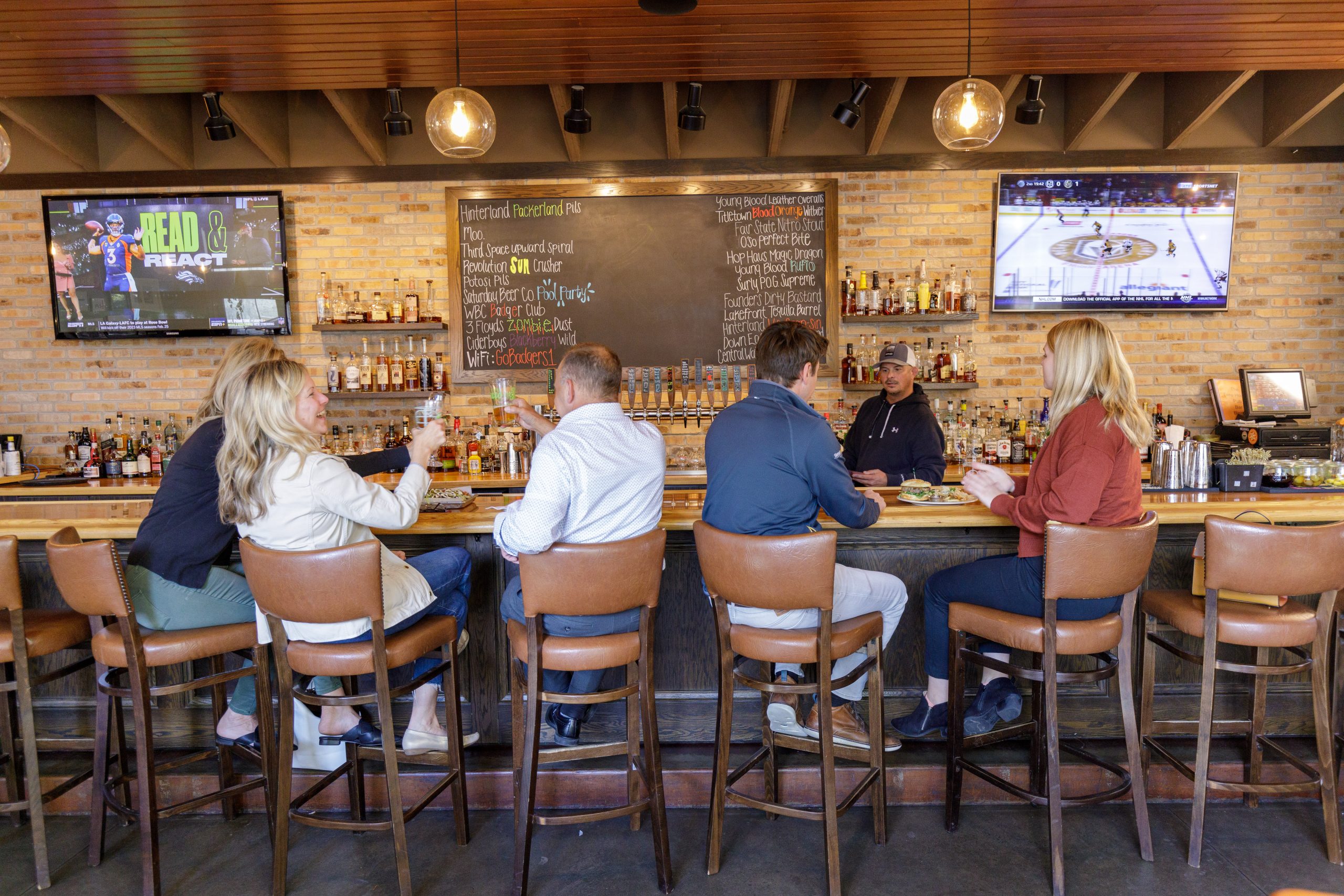 Patrons sit at a bar with two TVs displaying sports. A chalkboard lists drink options.