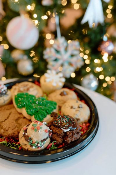 A tray of assorted holiday cookies with festive decorations is set on a table in front of a Christmas tree with ornaments and lights.