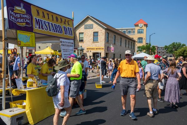 People attend an outdoor event for National Mustard Day, with a booth and signs for the Mustard Museum visible among the crowd on a sunny day.