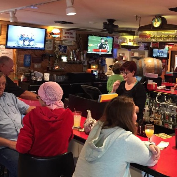 People sit at a bar counter with drinks while a bartender works. TVs display football games, and various decorations and signs, including some featuring a badger, are visible on the walls.
