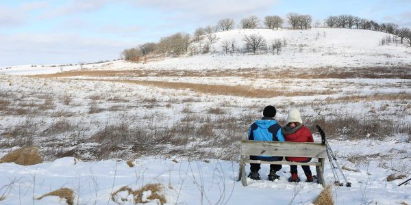 Two people in winter clothing sit on a bench overlooking a snowy field and distant hill under a cloudy sky.