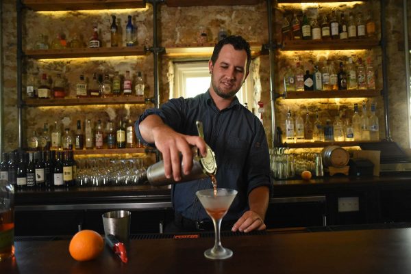 A bartender pours a cocktail from a shaker into a martini glass, expertly balancing the middle things of flavor and style, at a bar with shelves of bottles in the background.
