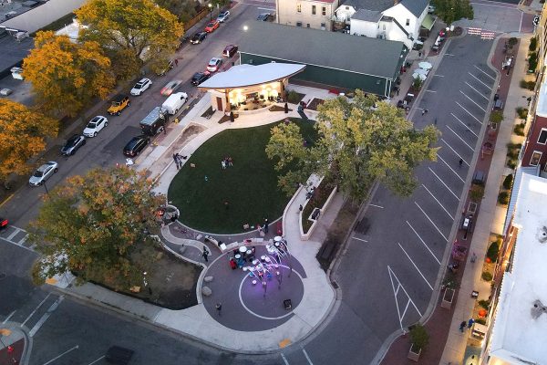 Aerial view of Stone Horse Green, a vibrant green space featuring a lush lawn, surrounding trees, curved sidewalk, empty parking spaces, and a few people gathered near a circular area with lights.