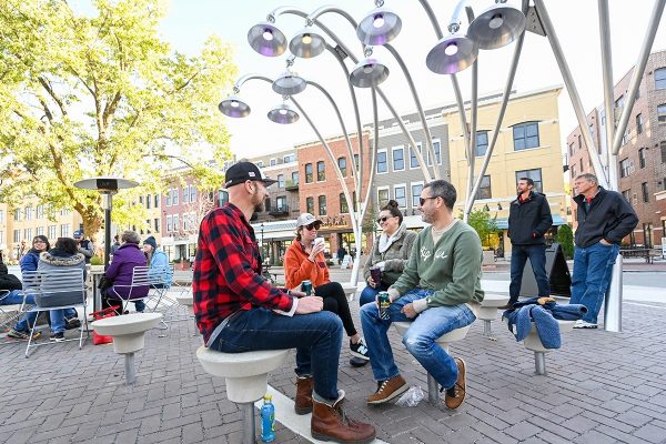 A group of people sit and talk on modern circular benches under metallic lamp structures at Stone Horse Green, an inviting green space in an outdoor urban plaza with buildings in the background.