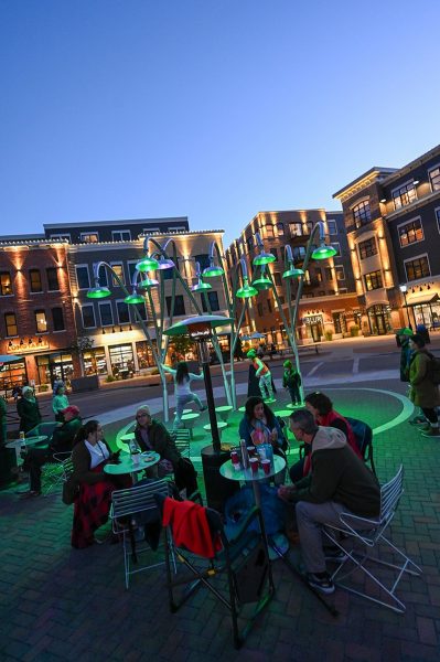 People sit and socialize at tables under green streetlights in Stone Horse Green, a vibrant outdoor plaza and green space, with brick buildings and shops in the background at dusk.