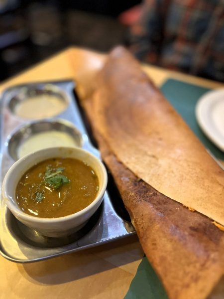 A crispy dosa served on a metal tray with three small bowls of sauces and chutneys on the side.