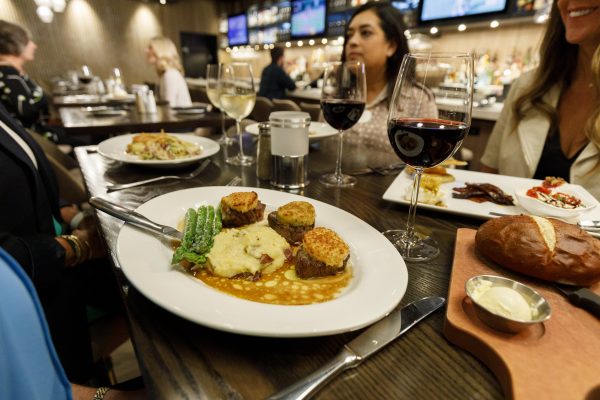 A plate with three pieces of meat, mashed potatoes, gravy, and asparagus is shown on a restaurant table. Two glasses of wine and other dishes are also visible. Four people are seated around the table.