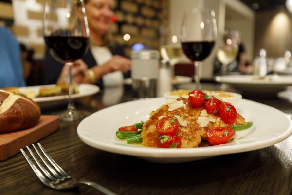 A plated dish of breaded chicken topped with cherry tomatoes and green beans sits on a table with glasses of red wine and other food items.