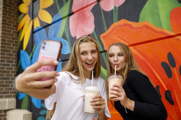 Two young women taking a selfie while holding milkshakes, standing in front of a colorful mural with flowers.