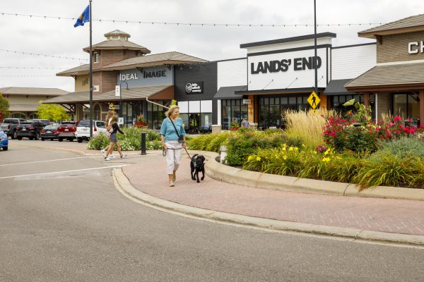 A woman walks a black dog on a leash along a paved pathway in a shopping center with storefronts like "LANDS' END" and "IDEAL IMAGE" visible in the background.