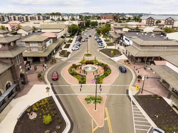 Aerial view of a shopping area with a roundabout, multiple stores, cars parked and driving, and a central American flagpole.