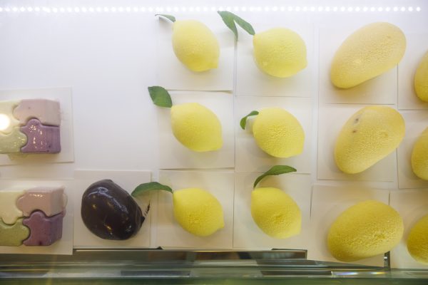 Display case with various desserts, including yellow lemon-shaped pastries, a dark oval pastry, and rectangular cakes in pastel colors.