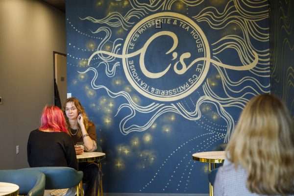 Two individuals sit at a table near a mural displaying "C's Patisserie & Tea House" in cursive on a blue background adorned with swirls and stars, while another person sits in the foreground.