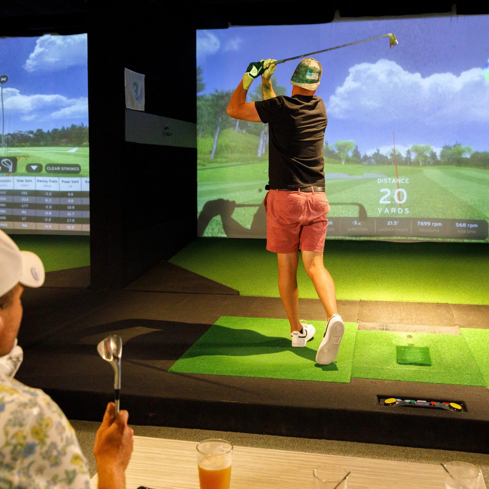 A person follows a Spring itinerary by swinging a golf club at an indoor golf simulator, while another watches from a table with drinks in the foreground.
