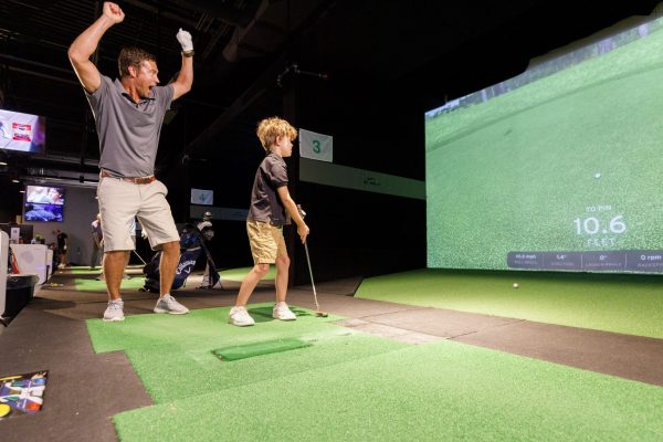 An adult celebrates while a child practices golf on an indoor putting green with a screen display.