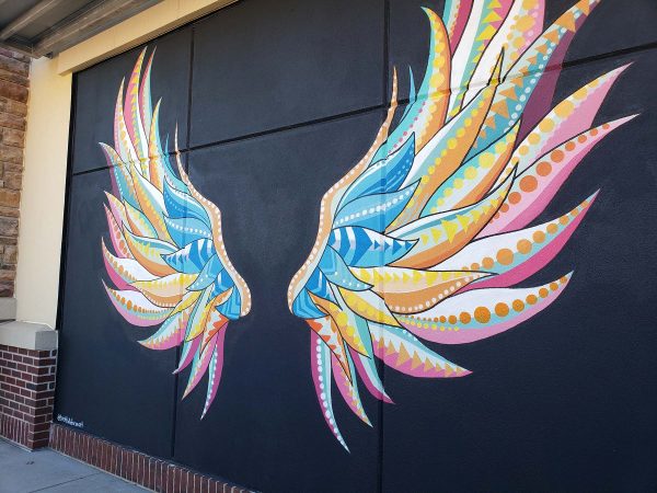 A vibrant mural of colorful, stylized wings is painted on a black wall, with shades of blue, yellow, orange, and pink forming feather patterns.