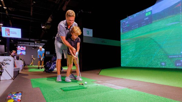 An adult helps a child practice golf using a simulator indoors; other people are seen using golf equipment in the background.