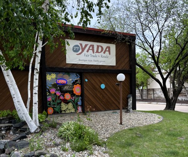 YADA Fair Trade & Resale shop with a sign above the entrance, a decorated window, and trees and rocks in the landscaped front yard.